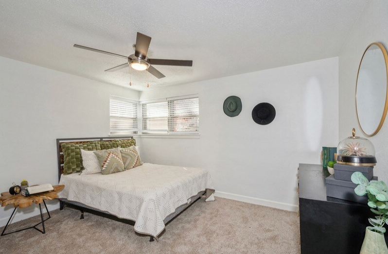 Bedroom with a bed, dresser, table, and mirror. White walls, dark bed frame, ceiling fan.