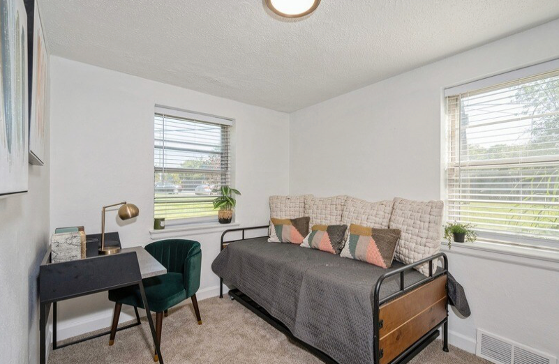 Small bedroom with daybed, desk, and two windows. White walls and neutral carpet.