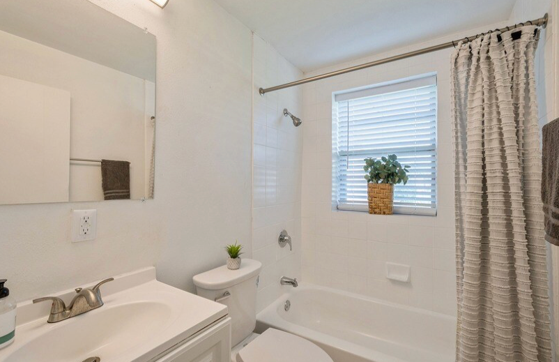 White bathroom with a sink, toilet, tub, and window with blinds.
