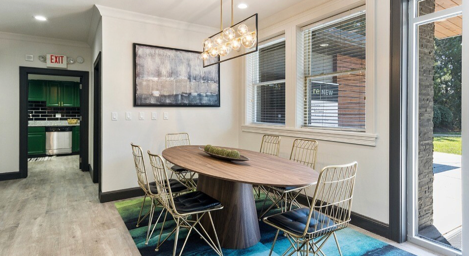 Dining room with oval table, gold chairs, art, windows, and chandelier. Door to kitchen visible.