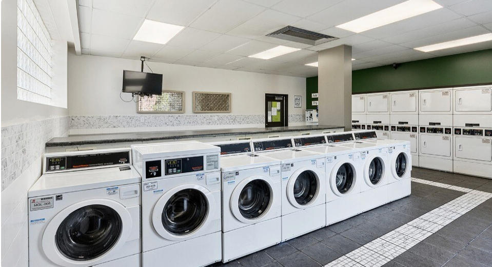 Row of washing machines in a laundry room with a countertop, TV, and stacked dryers.
