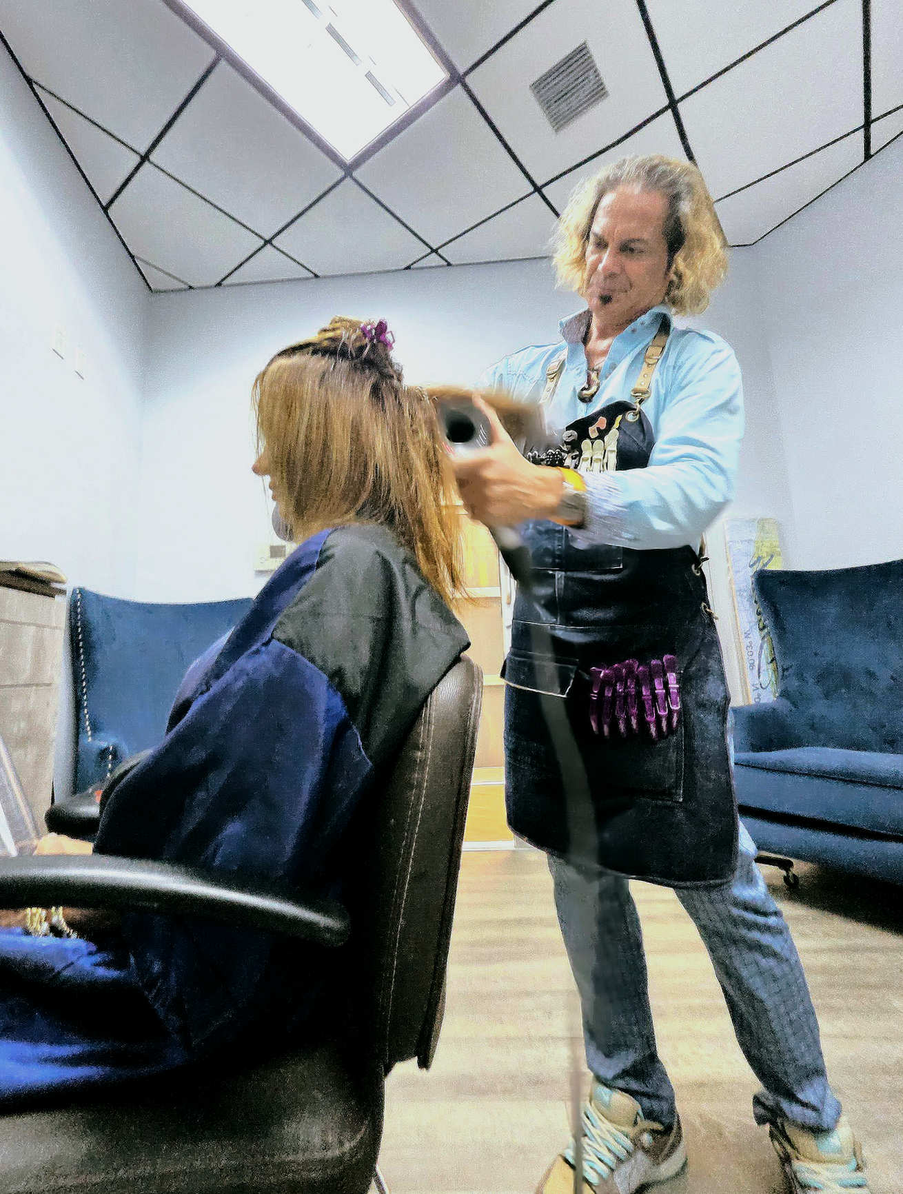 Hairdresser styling a client's hair in a salon. Blonde curly hair, blue shirt, black apron.