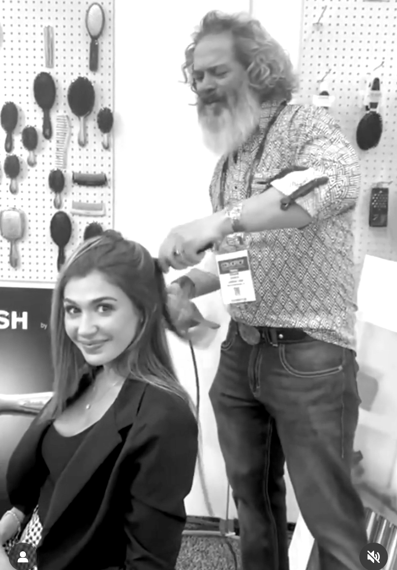 Woman having her hair styled by a man. They are indoors. Hairbrushes are displayed on the wall behind them.