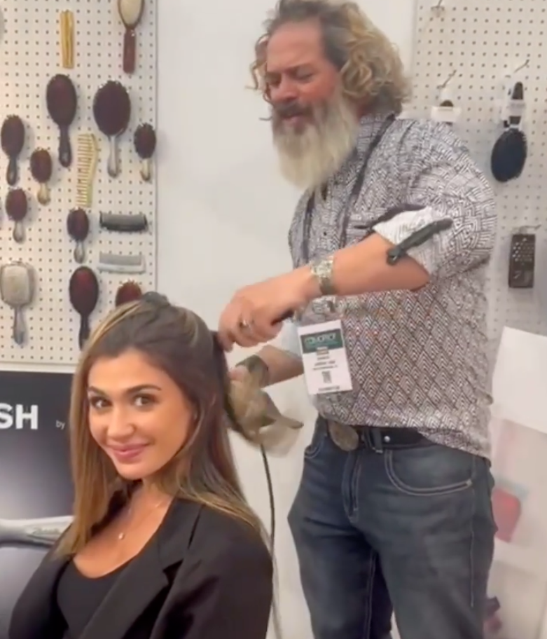A hairstylist uses a curling iron on a client's hair in a salon with various hairbrushes displayed on a wall.