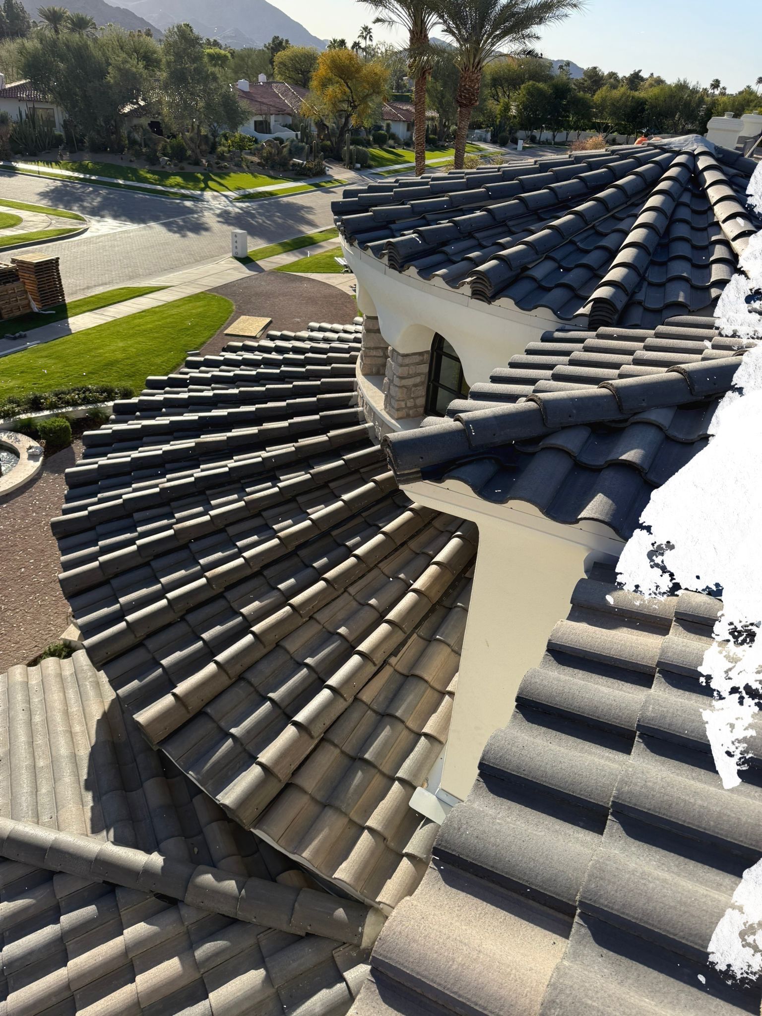 View of a tiled roof with curved sections, beige and dark gray tiles. Palm trees and a neighborhood are in the background.