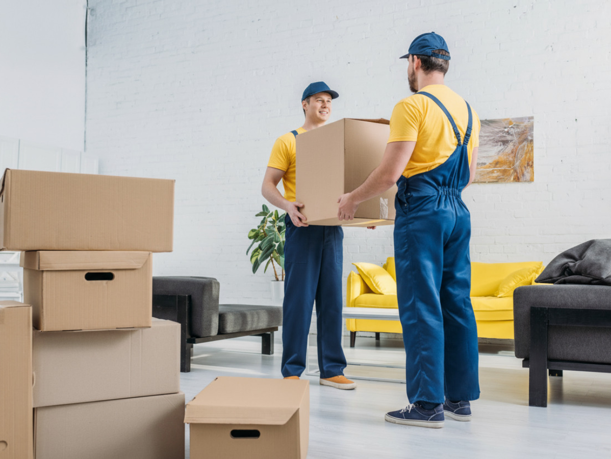 Two movers carrying a box in a living room with stacked boxes and a yellow couch