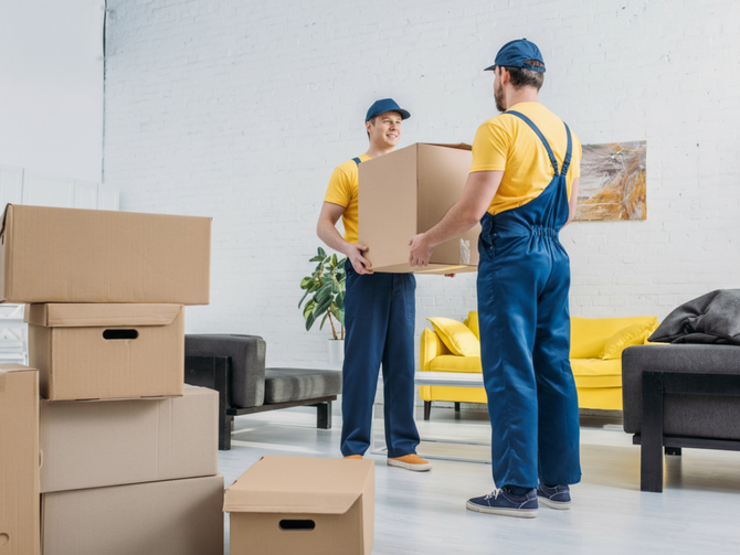 Two movers in yellow shirts and blue overalls carrying a box in a living room with stacked boxes