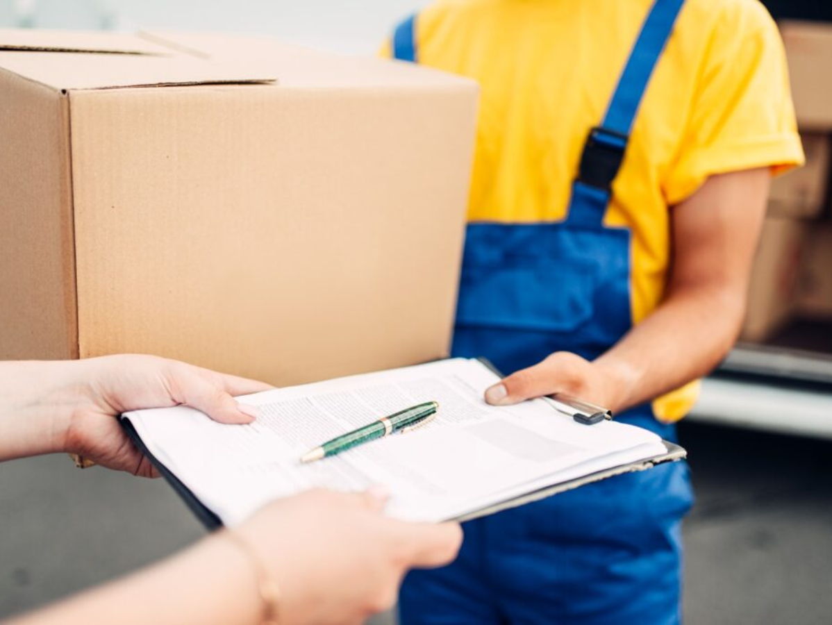 Delivery worker in blue overalls holding a clipboard beside cardboard boxes and a signed form