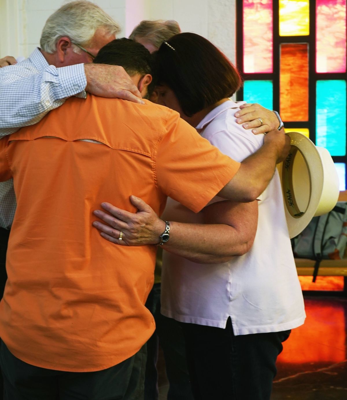 Three people in a church setting embrace with heads bowed in prayer, with stained-glass windows in the background.