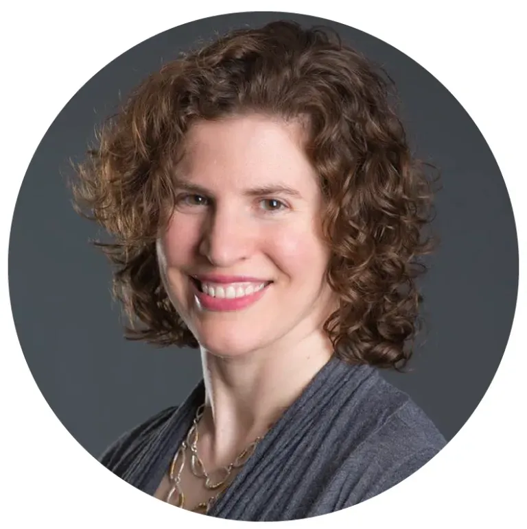Woman with curly brown hair smiles, wearing a gray shirt and gold necklace.
