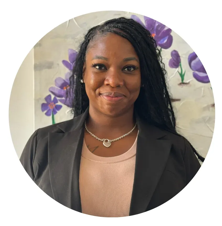 Woman with long dark braids wearing a blazer and necklace, smiling, in front of a painting of purple flowers.
