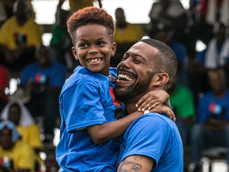 Man holding smiling child, both wearing blue shirts, laughing outdoors. Spectators in background.