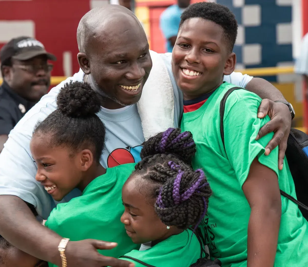 A smiling adult embraces three children, all wearing matching green shirts, in an outdoor setting.