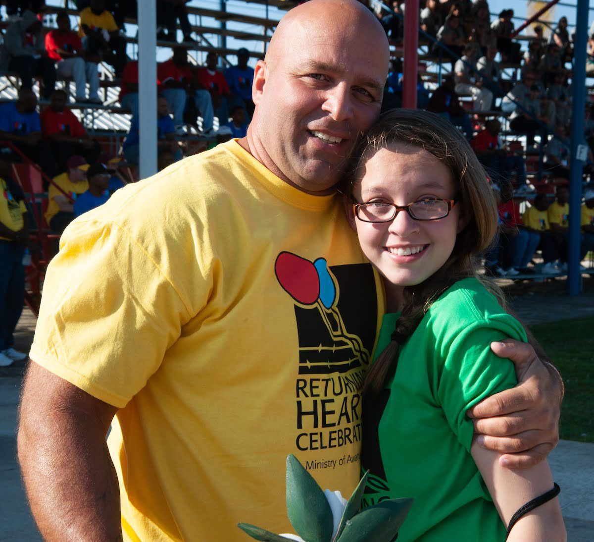A bald man in a yellow shirt hugs a smiling girl in a green shirt outdoors in front of stadium bleachers.