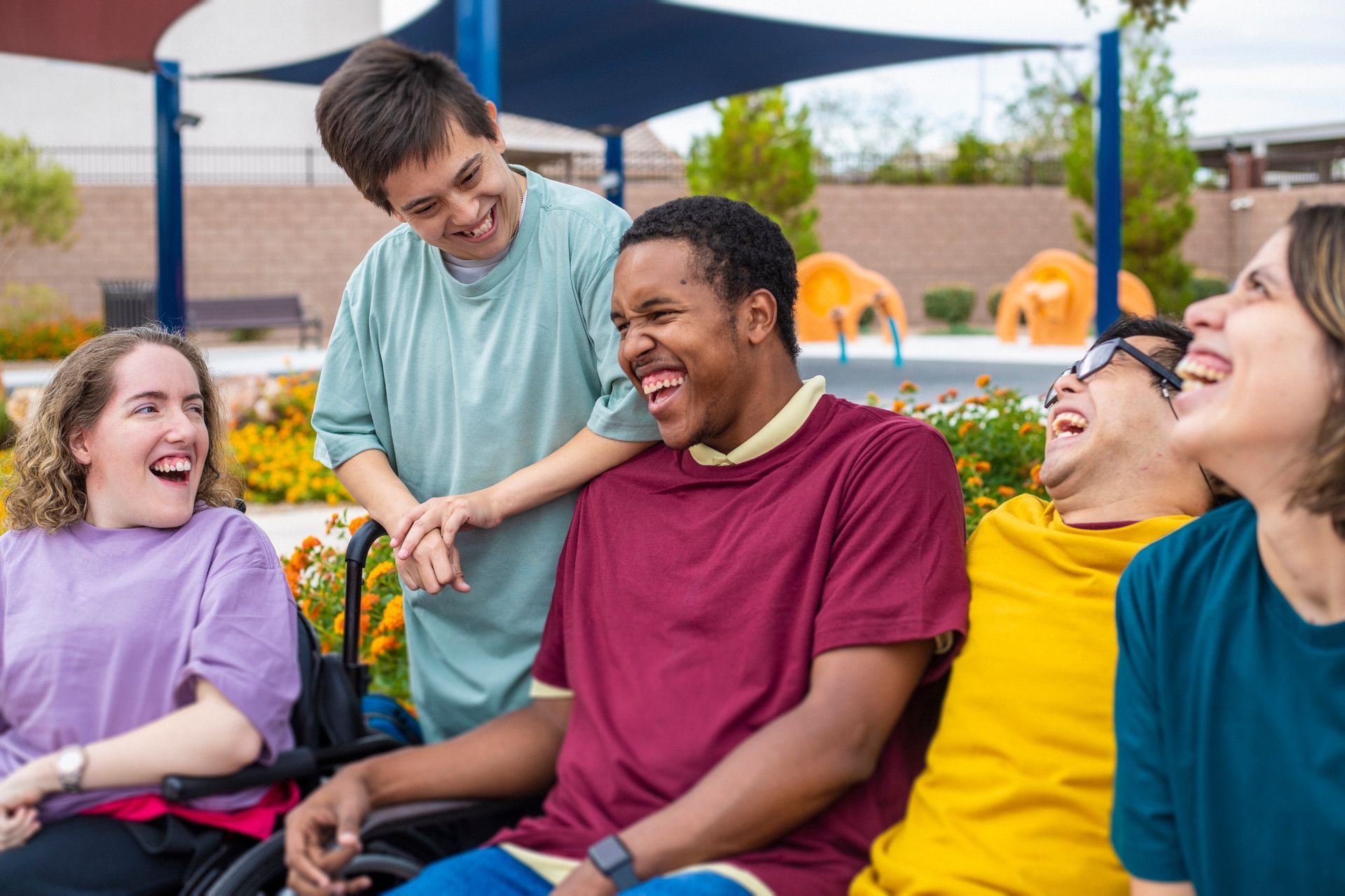 Group of people laughing outdoors. Some use wheelchairs. Flowers and blue canopy in background.