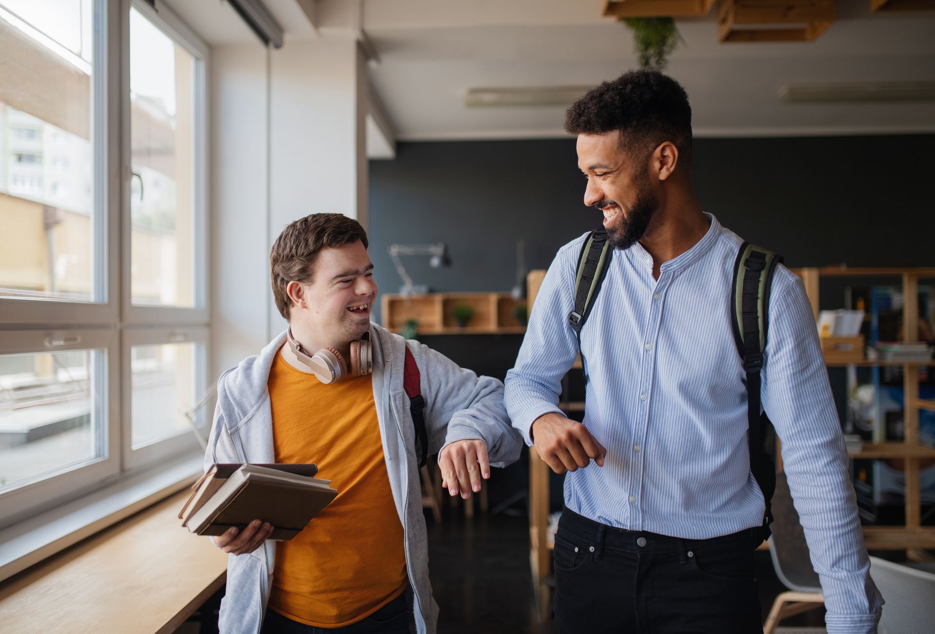 Two men greeting each other with elbow bumps in a bright, modern room. One man holding books.