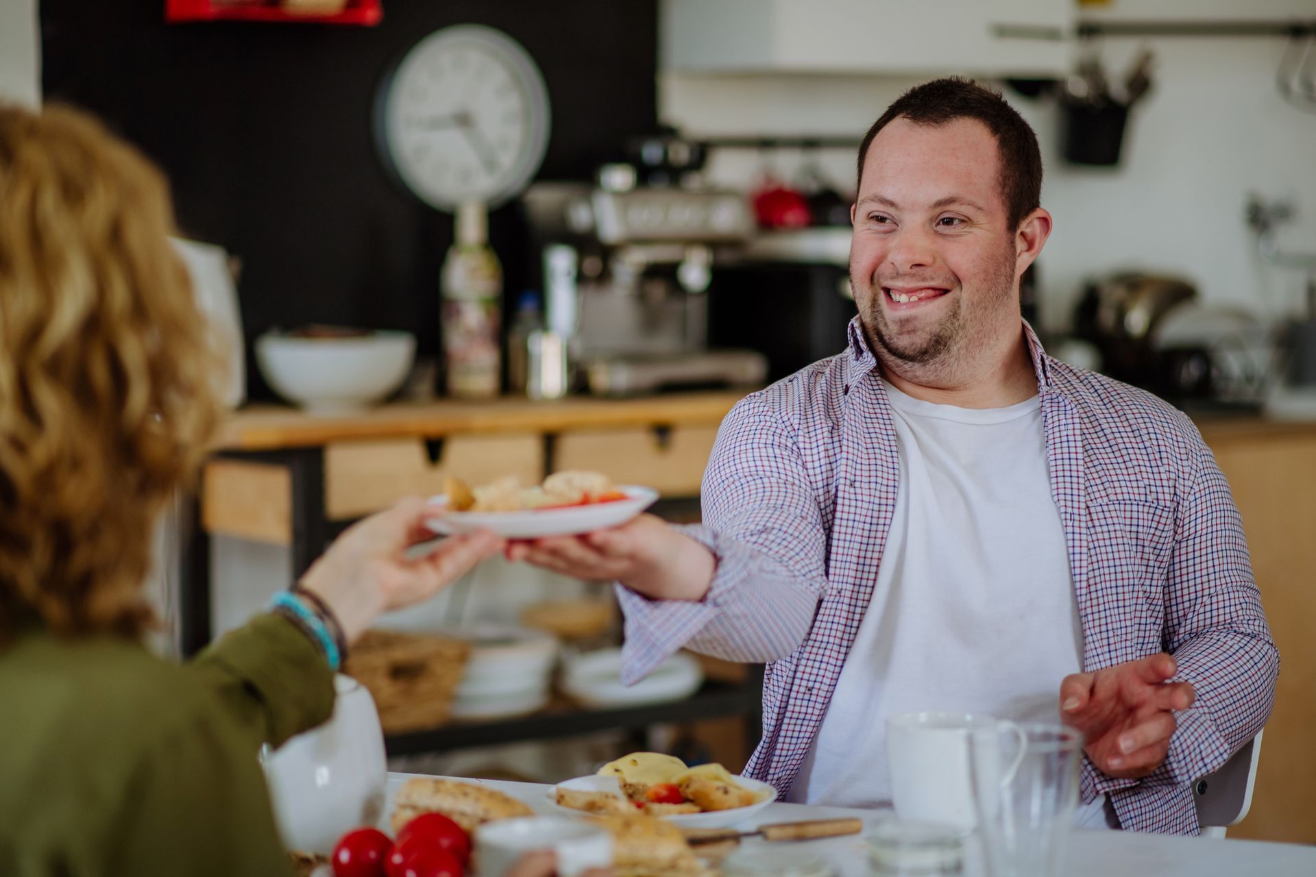 Man smiles, offers plate of food to person at a table, sunny kitchen setting.