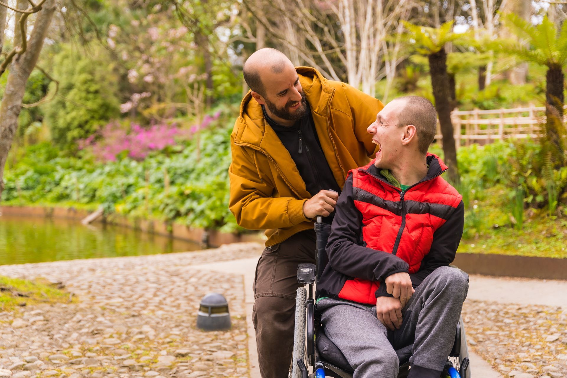 Man in yellow jacket pushing person in wheelchair, smiling and looking at each other outdoors, park setting.