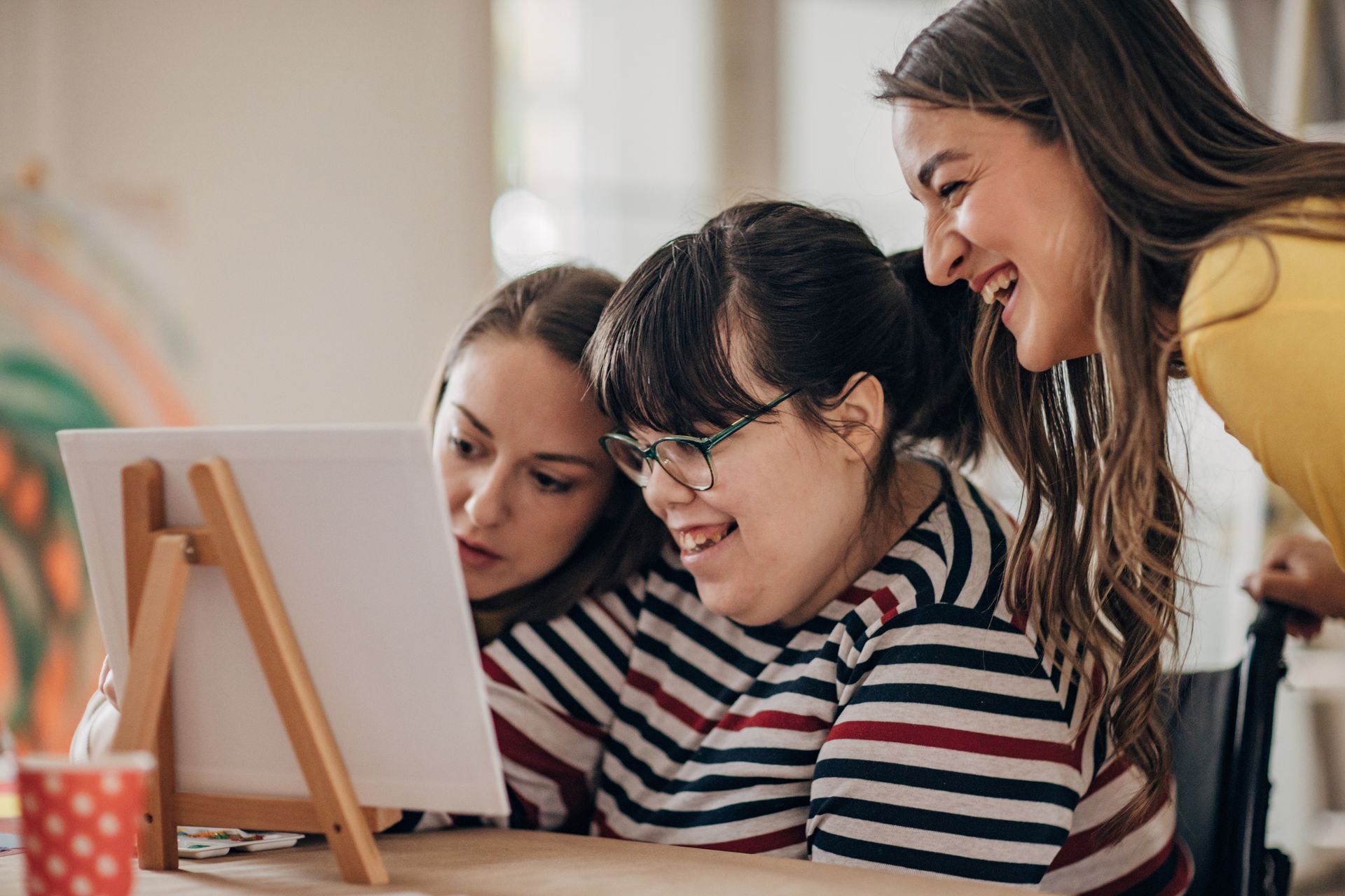 Three people looking at a painting on an easel. Two wear striped shirts, and one smiles broadly.