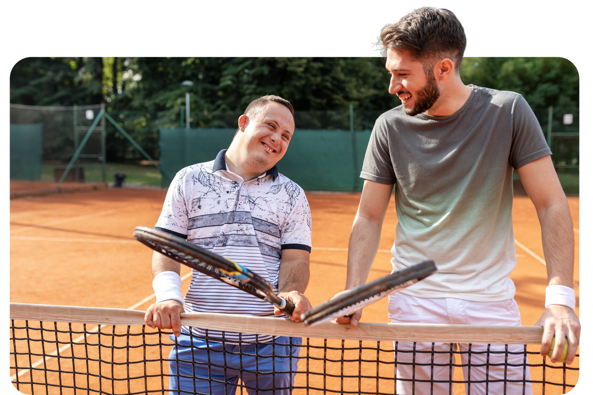 Two men, one with Down syndrome, smile and hold tennis rackets by a net on a clay court.