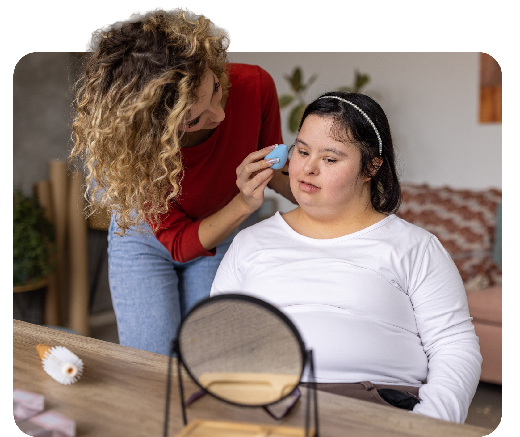 Woman applying makeup to another woman's face, using a blue sponge; mirror and makeup on a table.