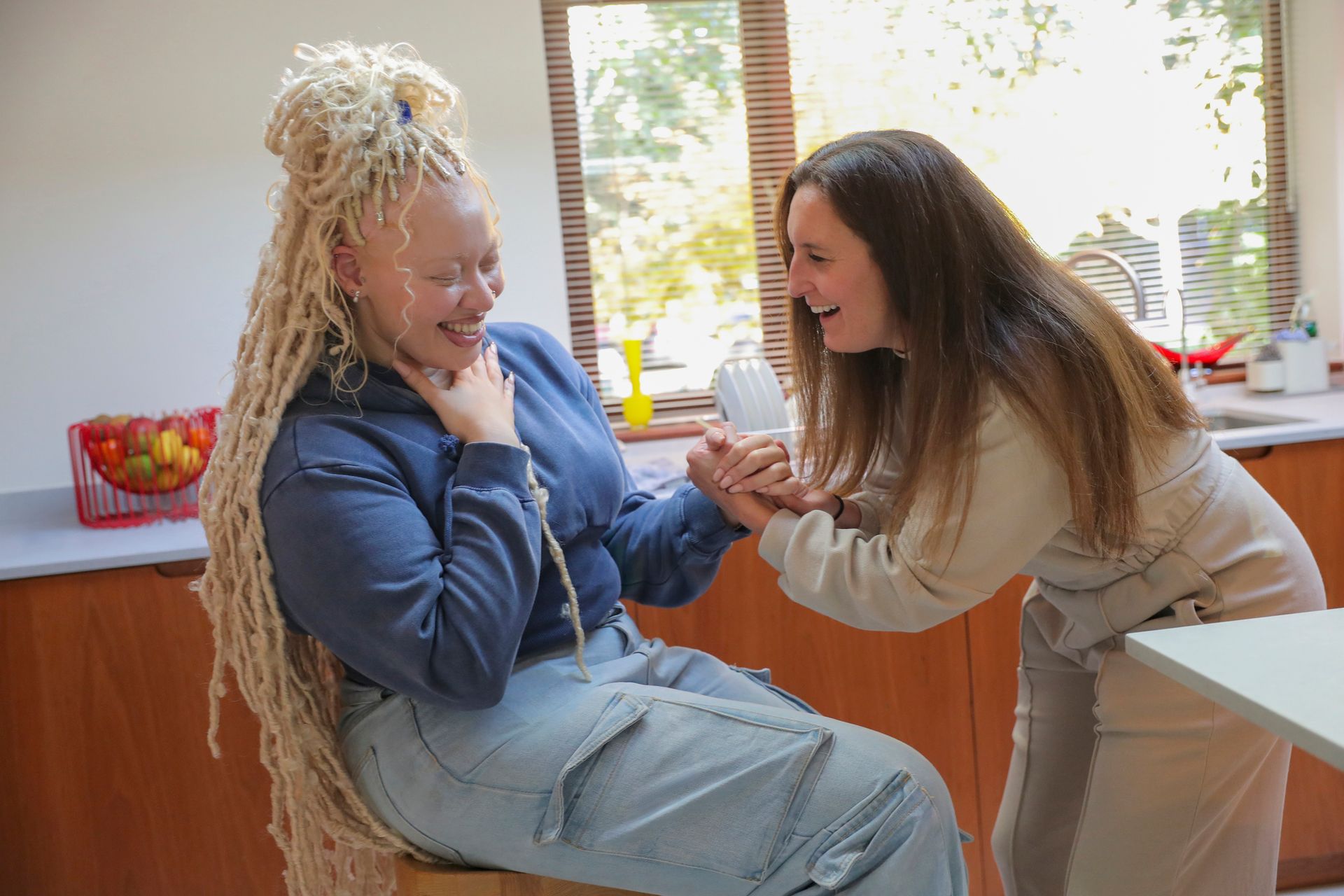 Two people laughing in a kitchen. One with long blonde dreadlocks, the other with brown hair, holding hands.