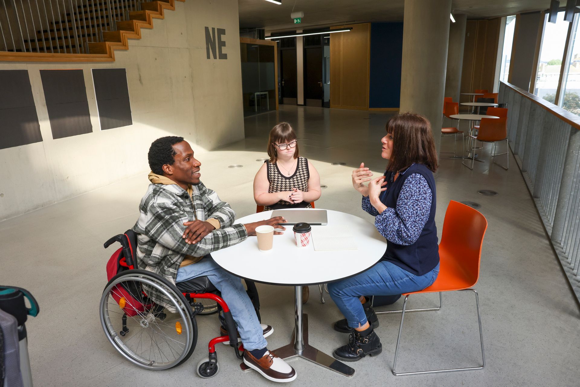Three people at a round table, one in a wheelchair, two others seated. Talking near a bright window.