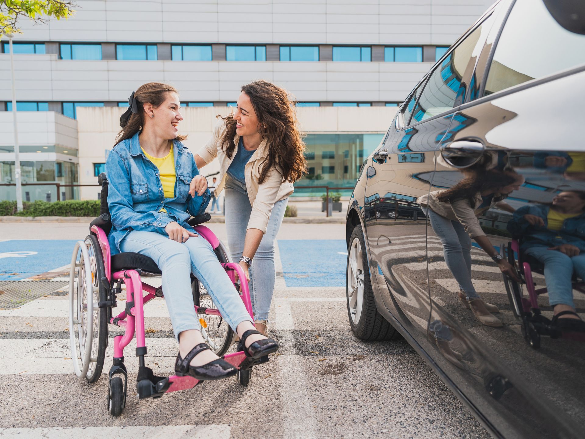 Woman in wheelchair smiling, helped by another woman at a car, near a building.