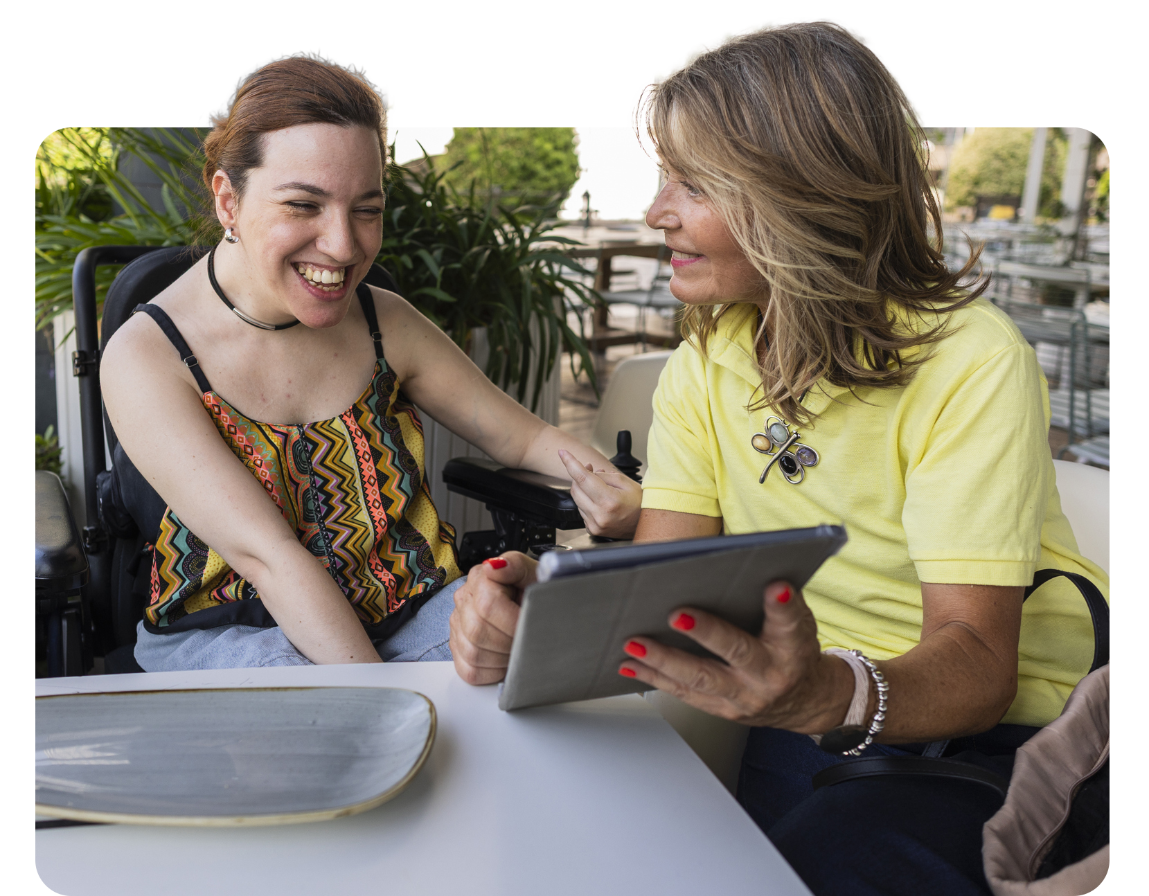 Woman in wheelchair smiles while another woman shows her a tablet at an outdoor table.