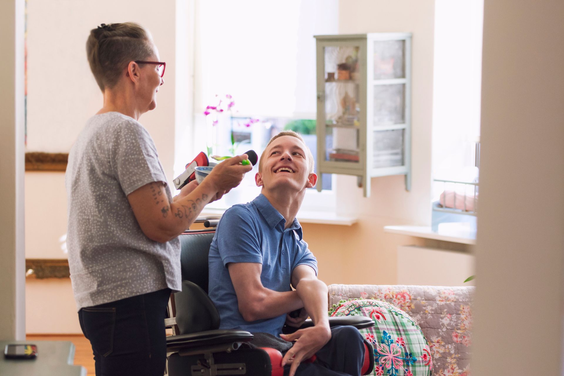 Woman holding a colorful toy for a man in a wheelchair; he smiles, looking up. Interior shot.