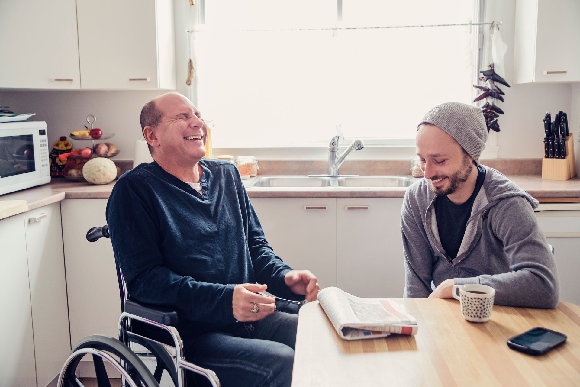 Man in wheelchair laughs with another man at kitchen table.