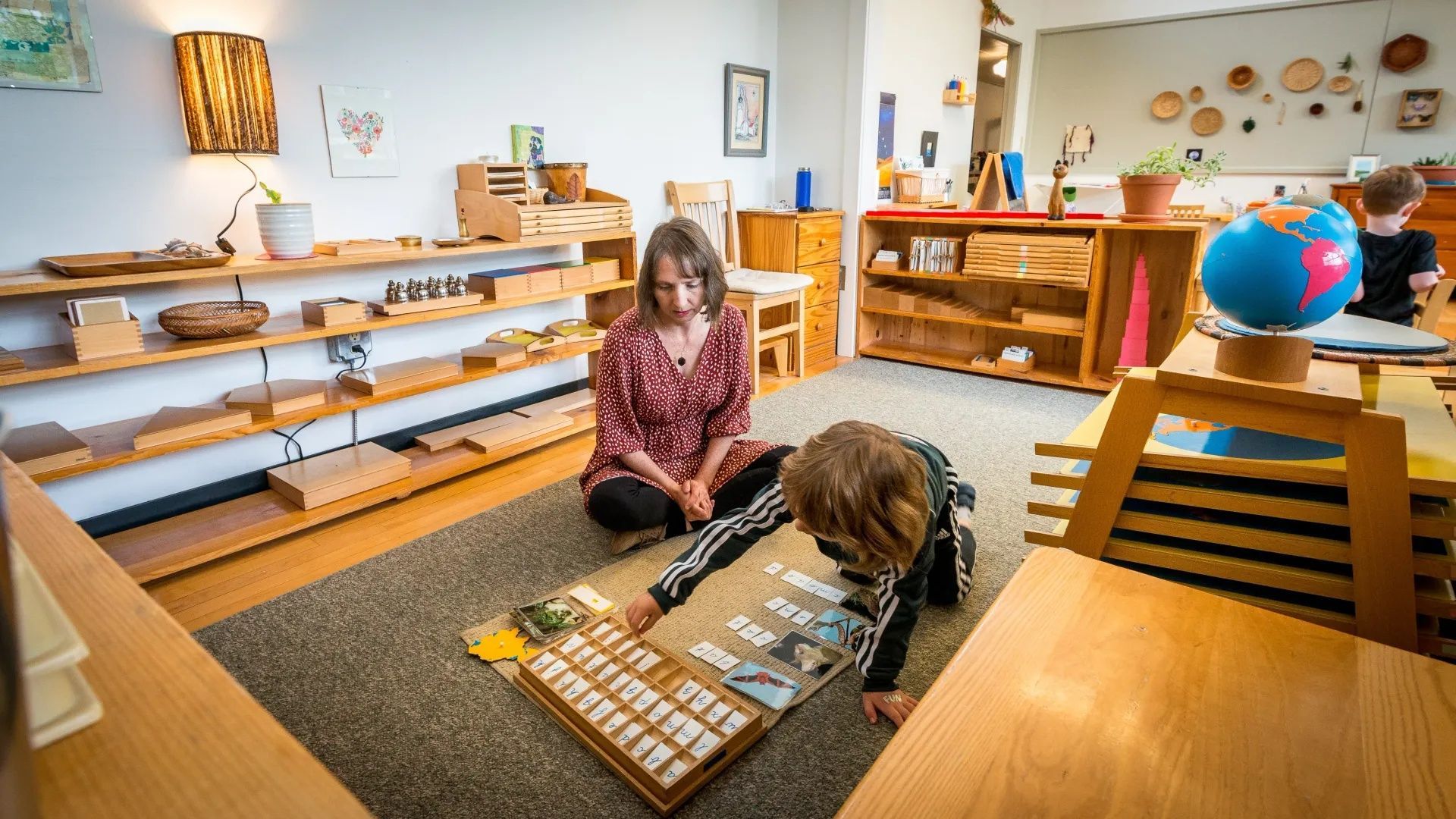 A Montessori Guide sits on the floor with a young child doing an educational activity.