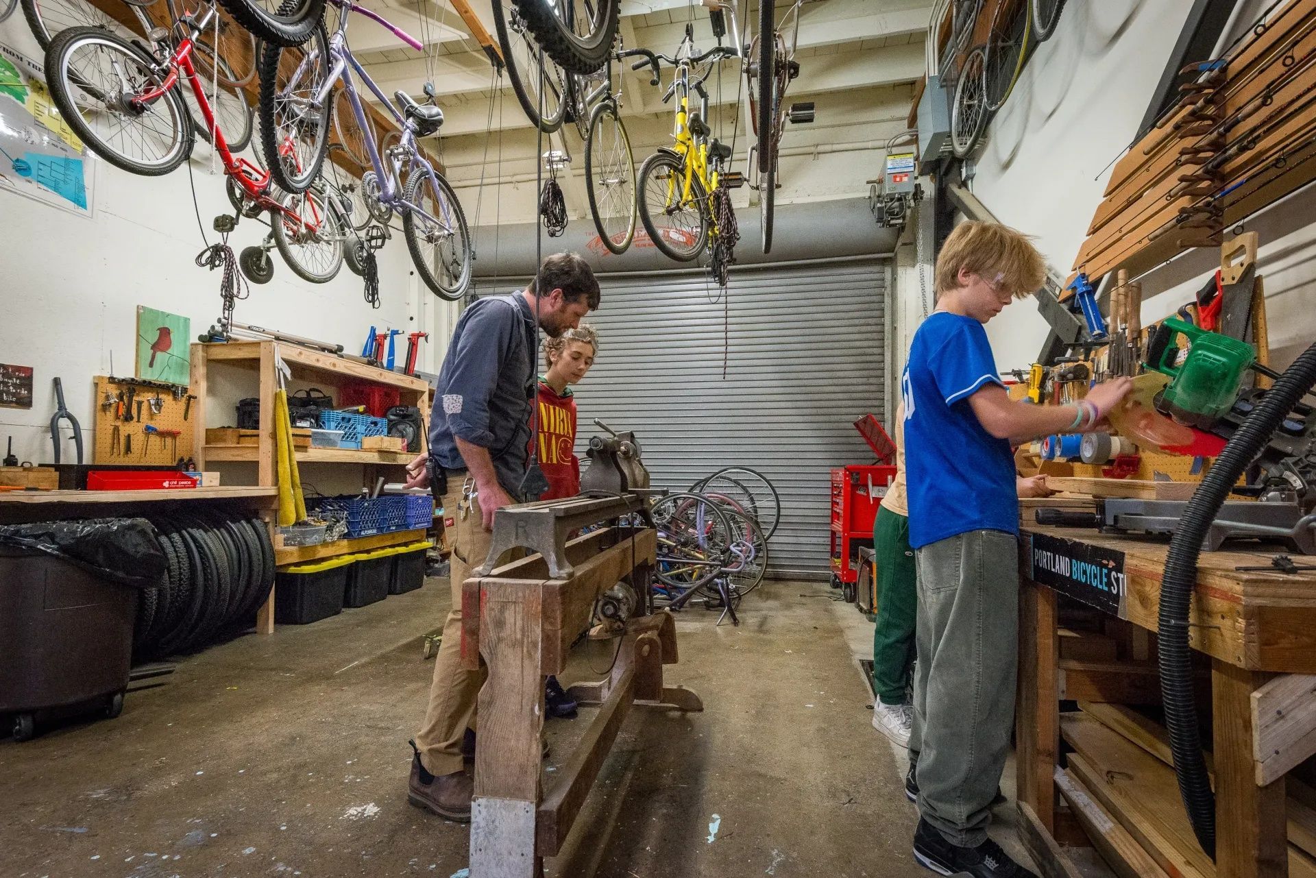 An adult and two middle school kids with blonde hair working in wood and bike workshop.