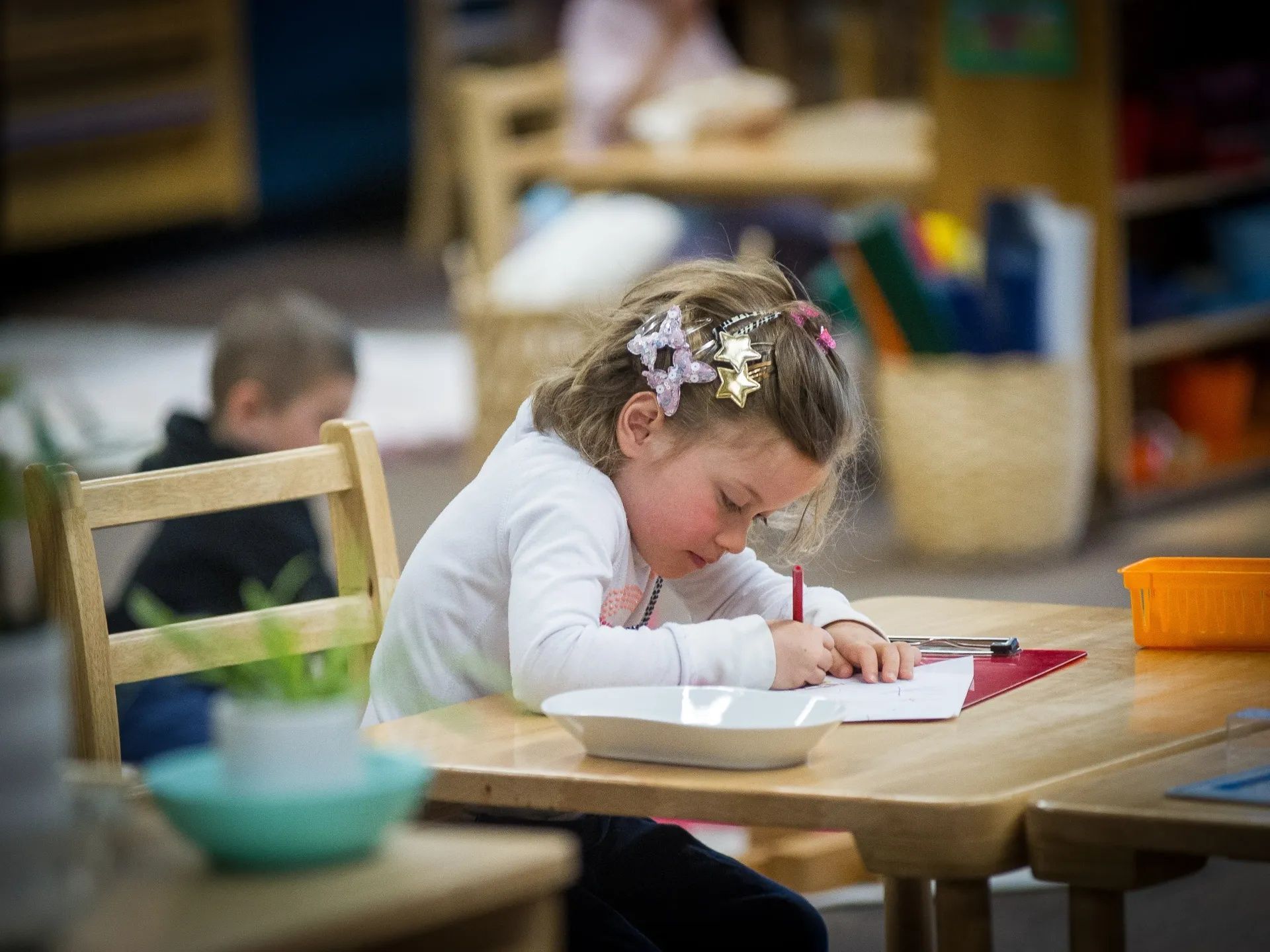 Young girl with blonde hair sitting at a wood table, writing with red marker.