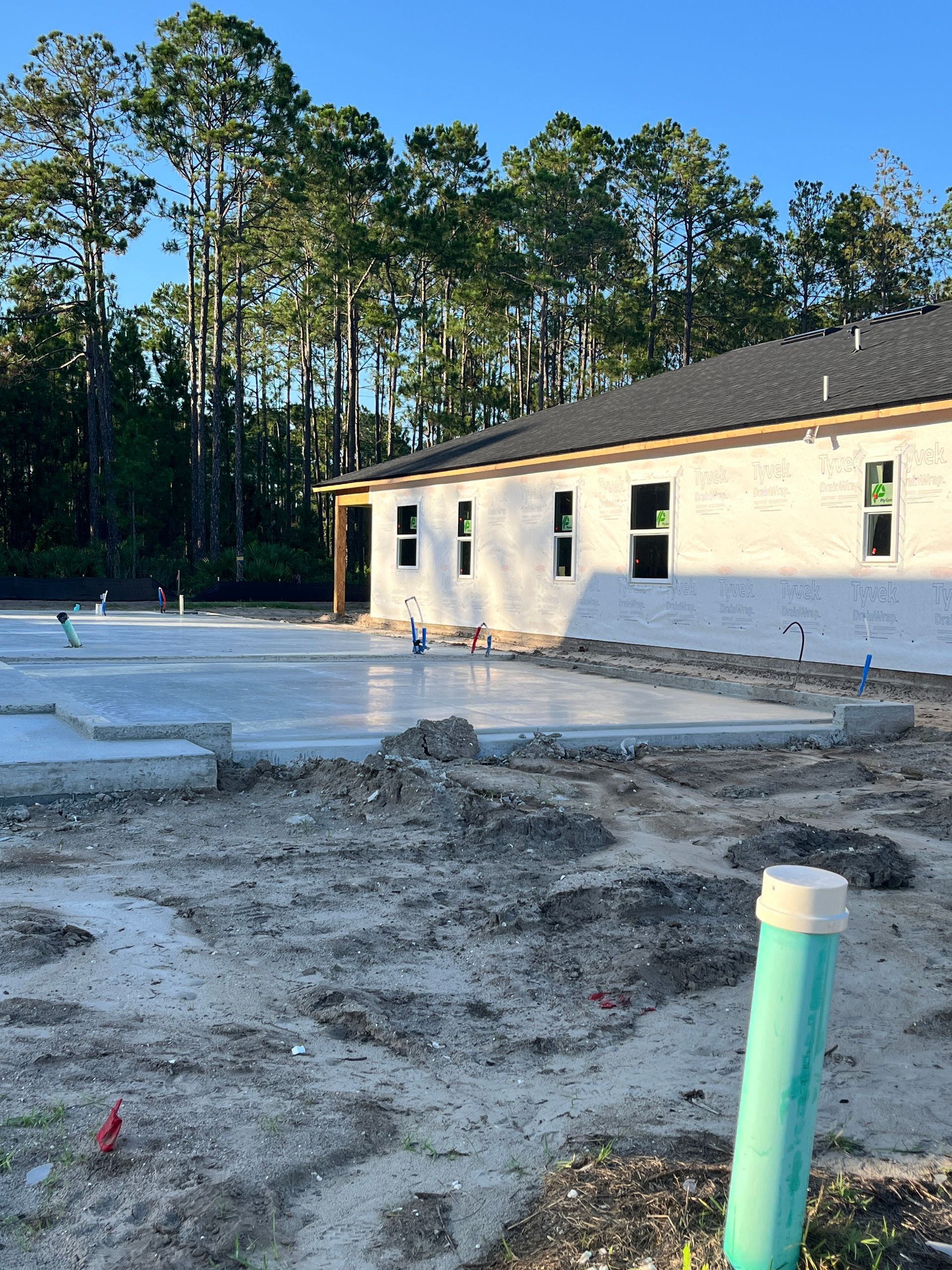 Construction site, concrete foundation, white brick walls, windows, green PVC pipe, trees in background.