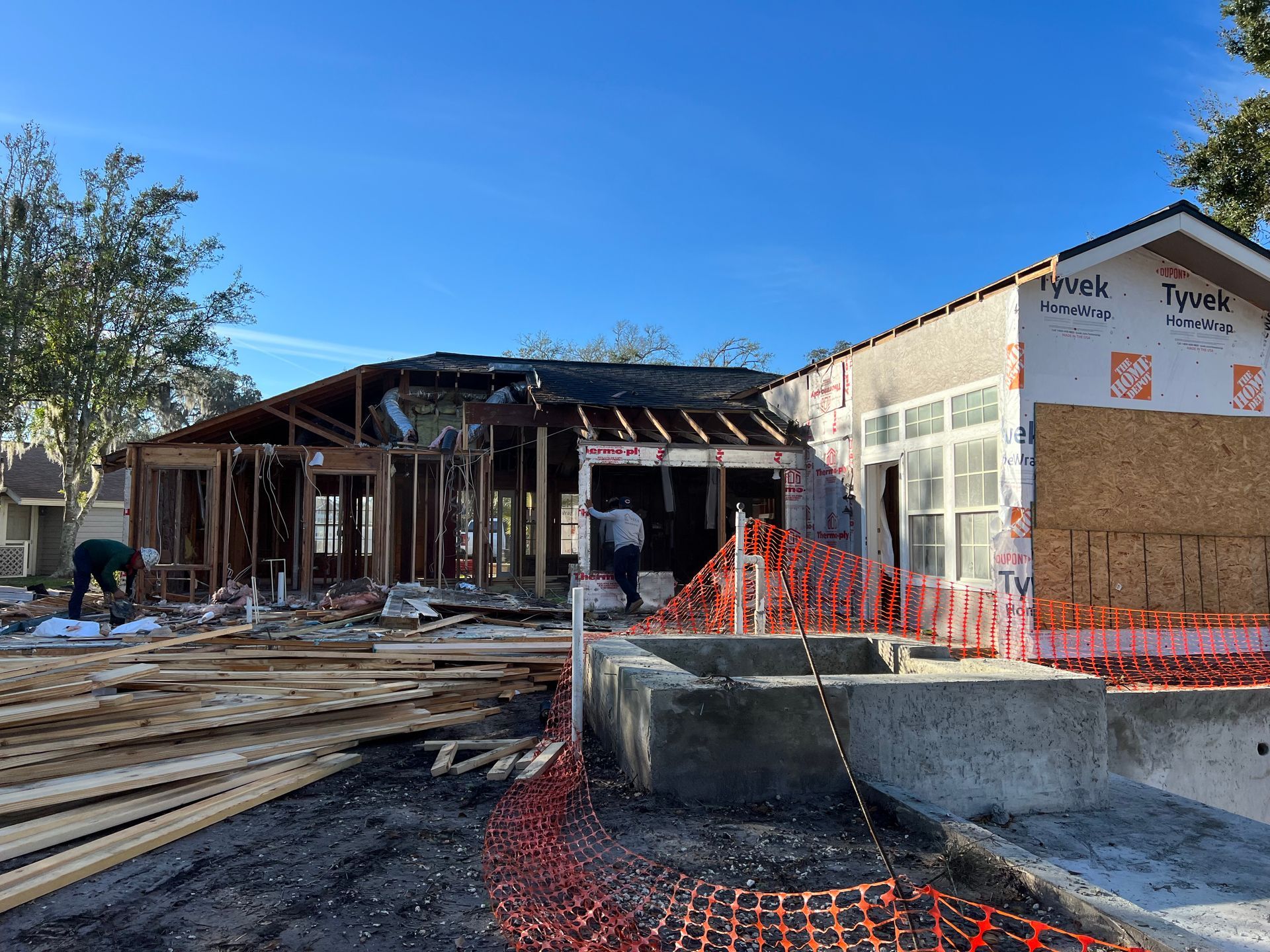 House under construction; wood framing exposed, workers present, surrounded by orange safety fencing, sunny outdoor setting.