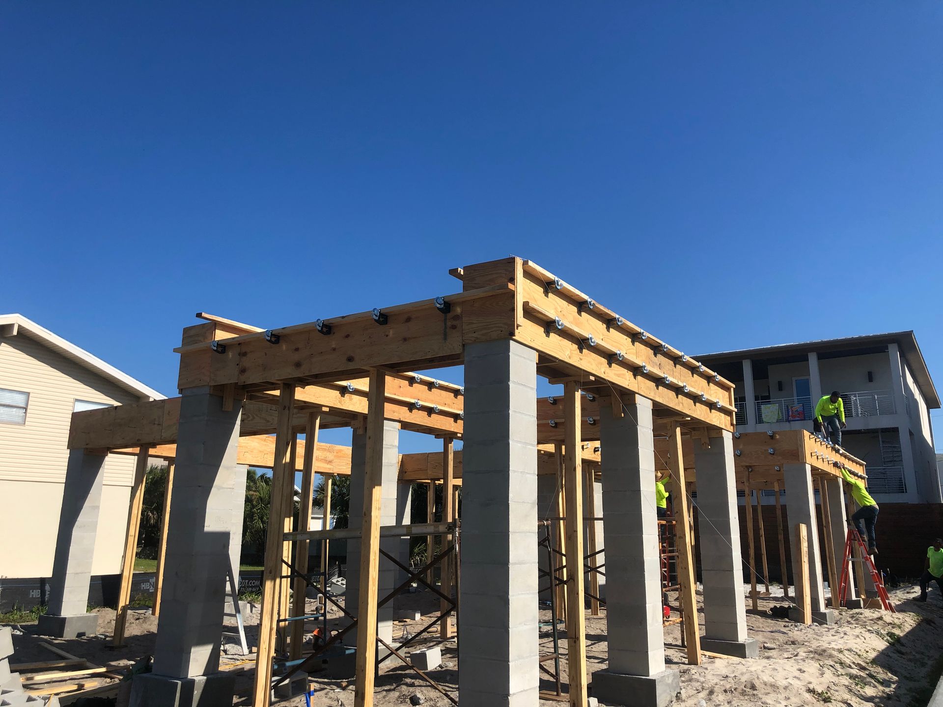 Construction site with concrete columns and wooden beams under a clear blue sky. Workers visible.