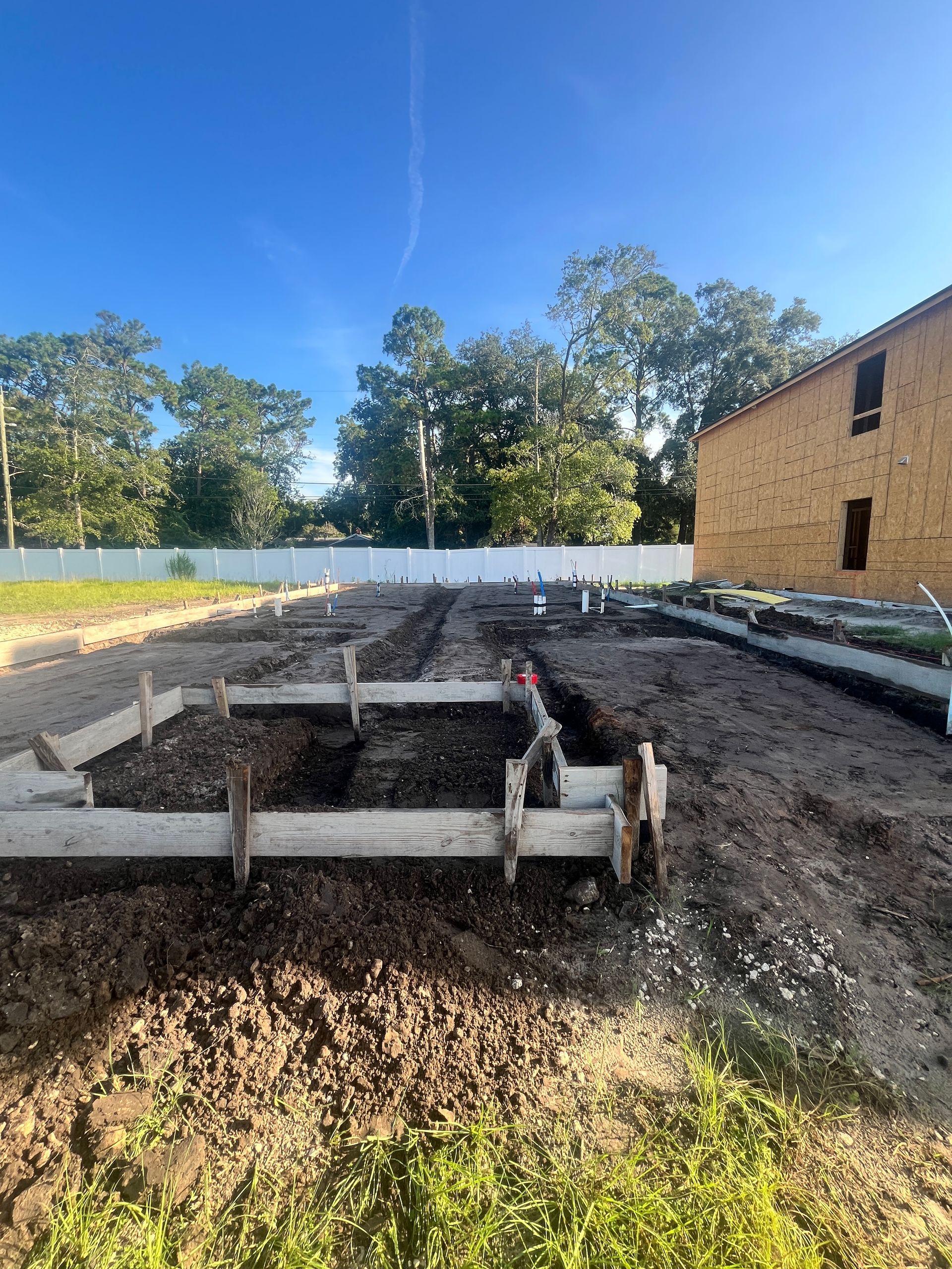 Construction site: wooden forms for foundation set in dirt, trees and building in the background.