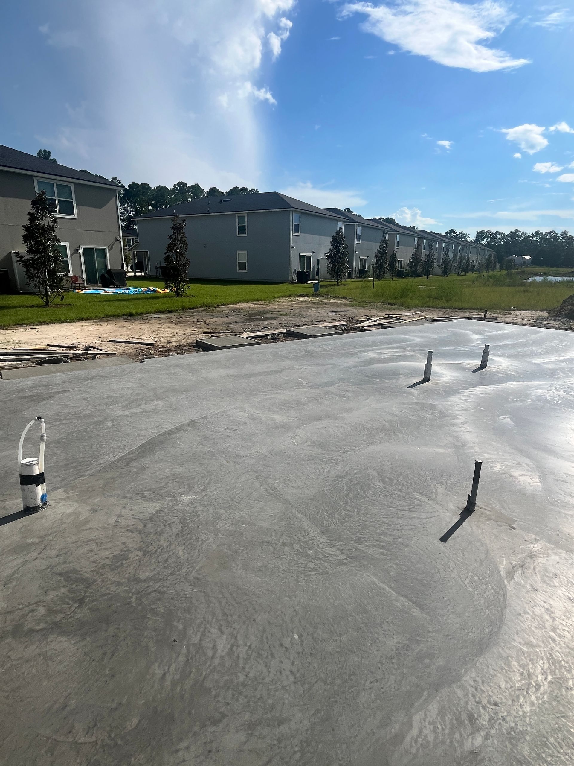 Newly poured concrete slab with plumbing fixtures, background of suburban houses and blue sky.