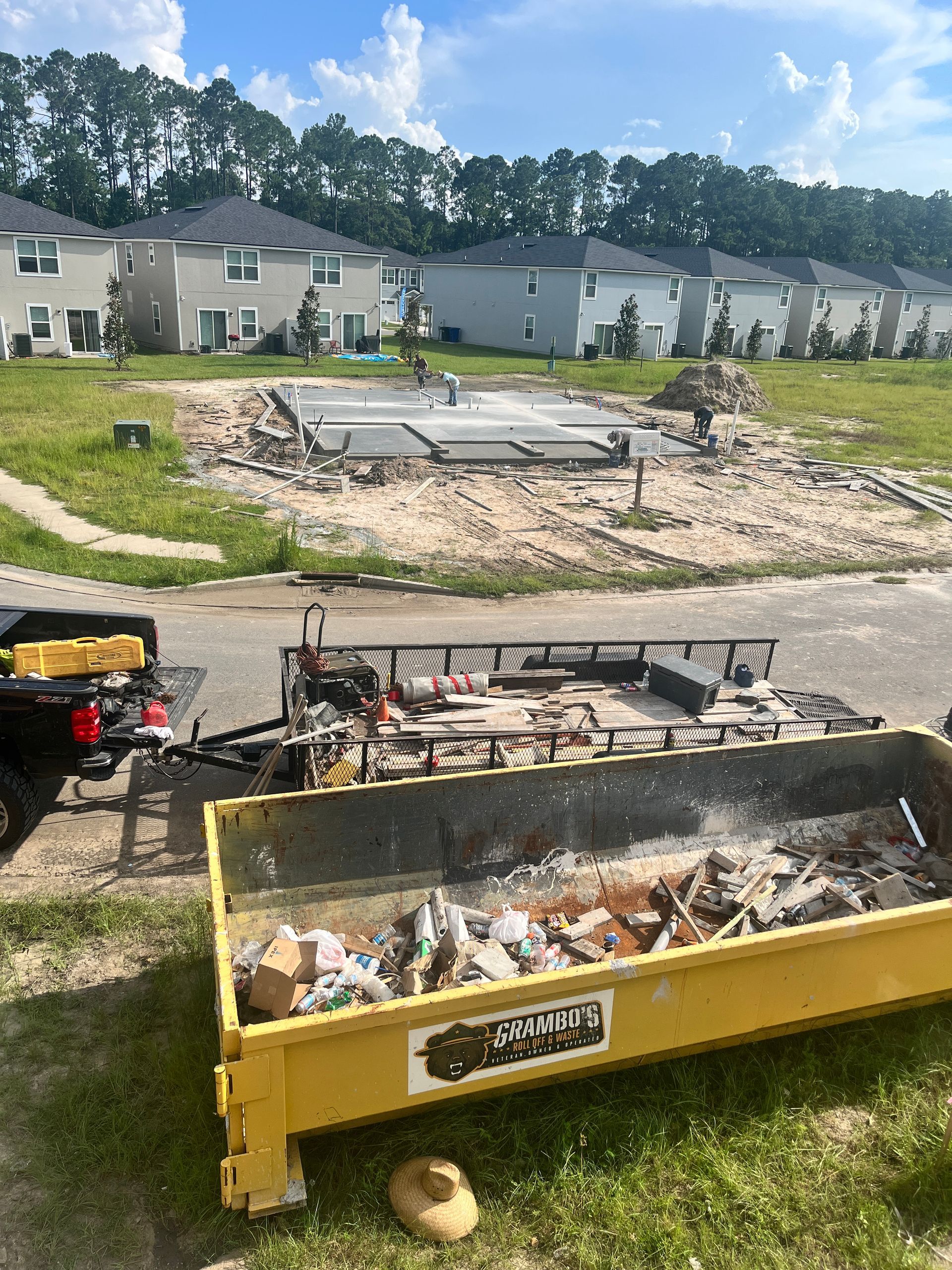 Construction site with a yellow dumpster, a trailer, and a concrete foundation. Houses in the background.