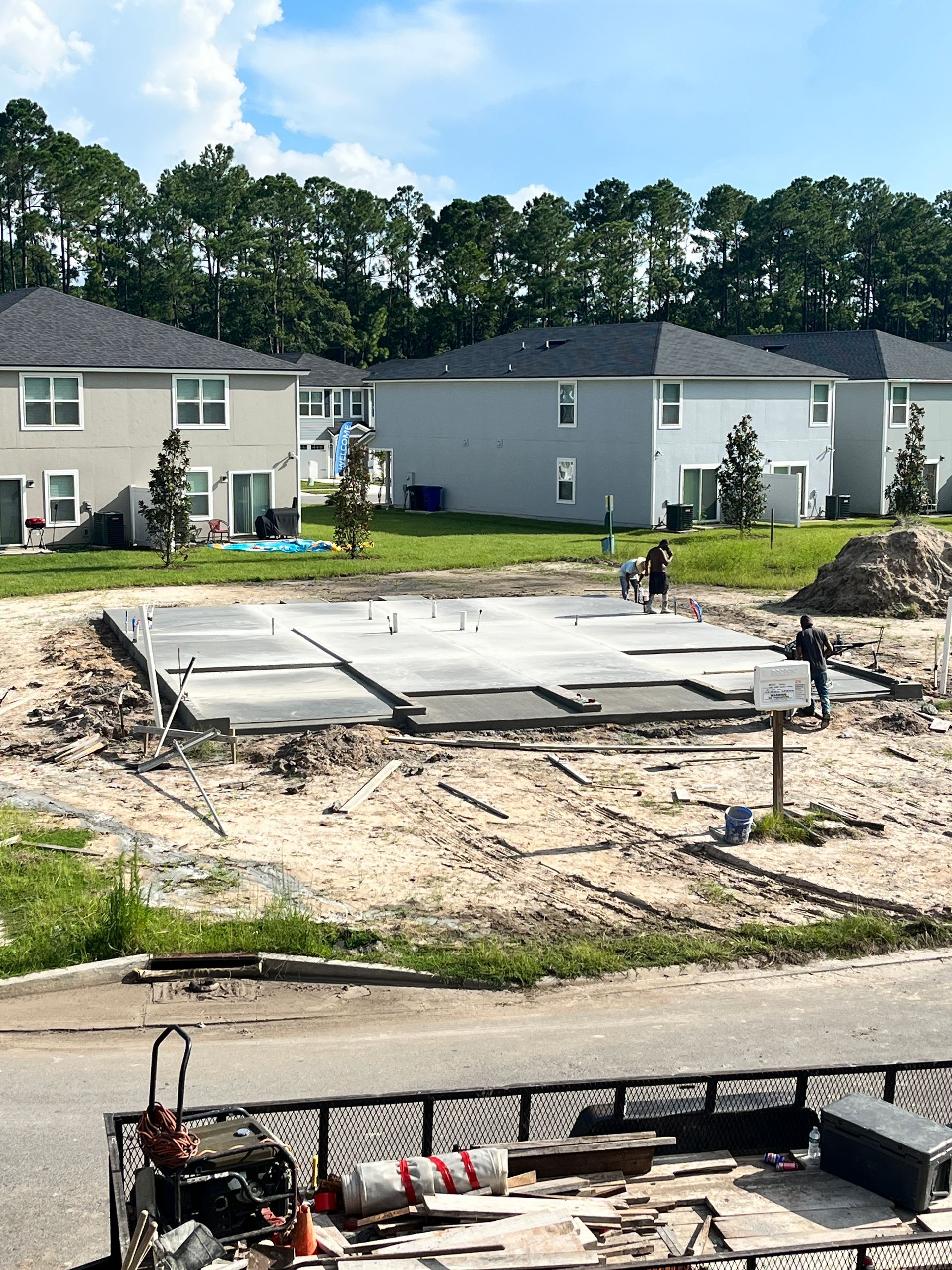 Construction site: concrete foundation with workers; houses in background; blue sky.