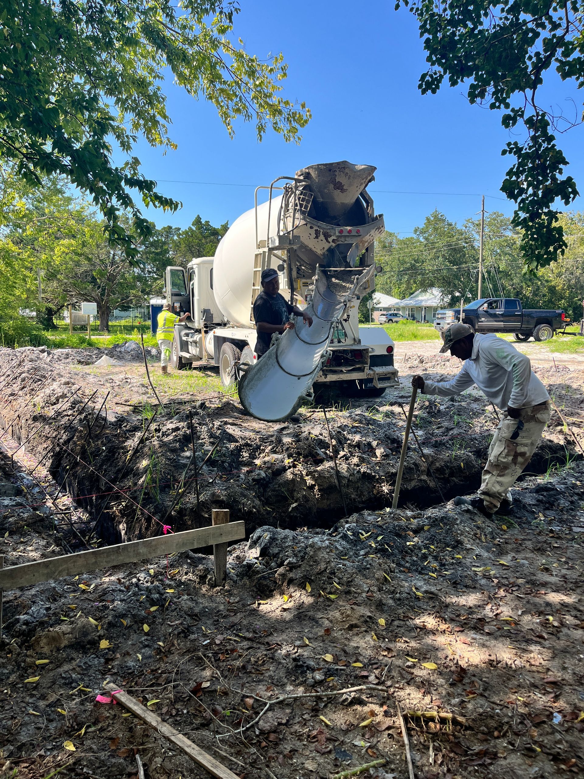 A concrete truck pours wet cement into an excavated foundation pit, as construction workers guide the flow on a sunny day.