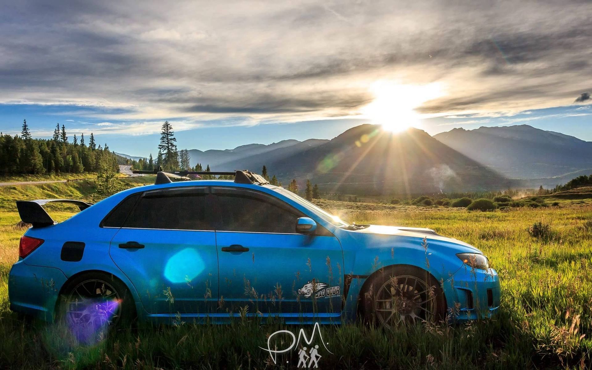 Blue Subaru sedan parked in a grassy field, with mountains and a sunset in the background.