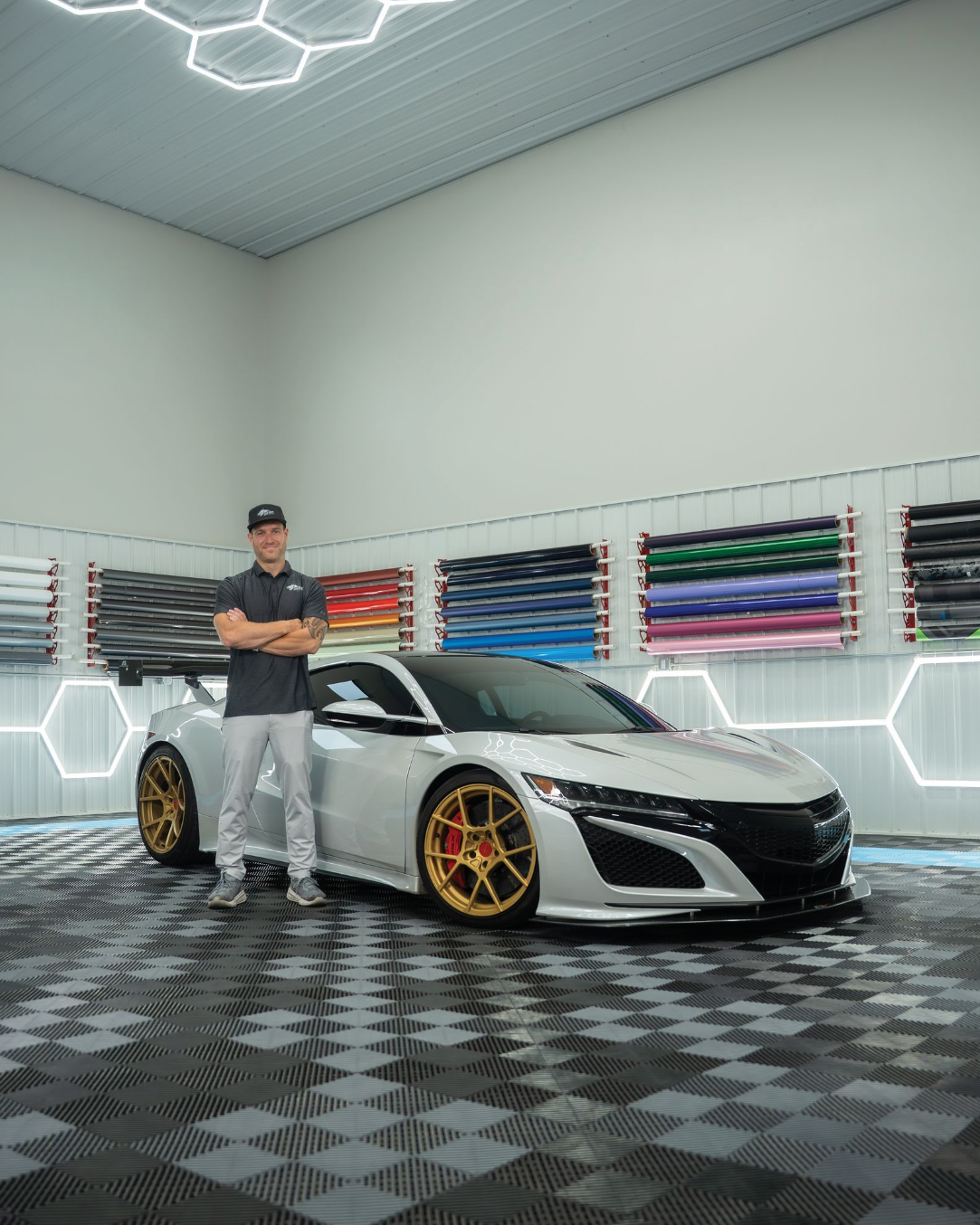 Man posing with a silver sports car, gold wheels, in a garage with color samples.