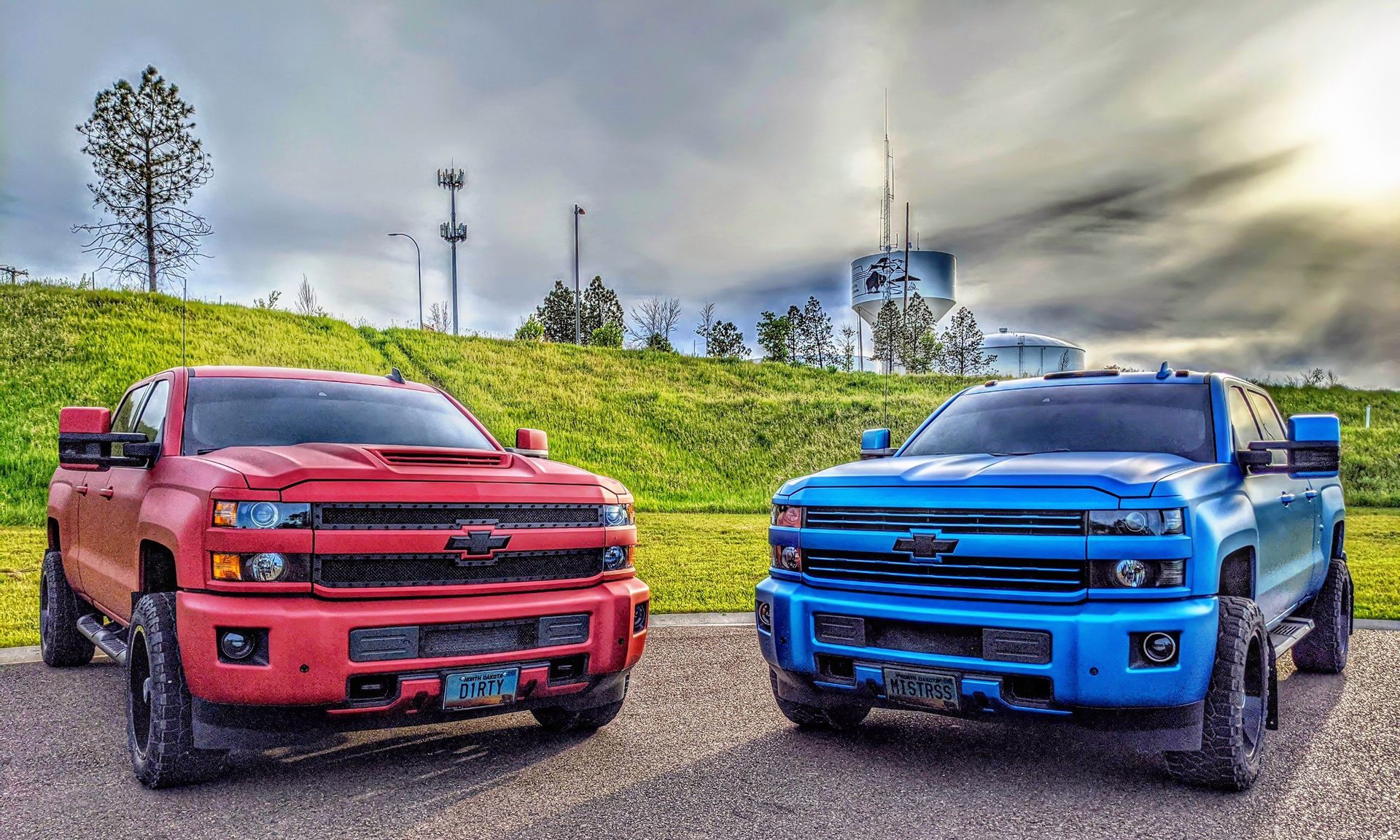 Two lifted red and blue Chevrolet trucks parked on pavement, green hill in background.