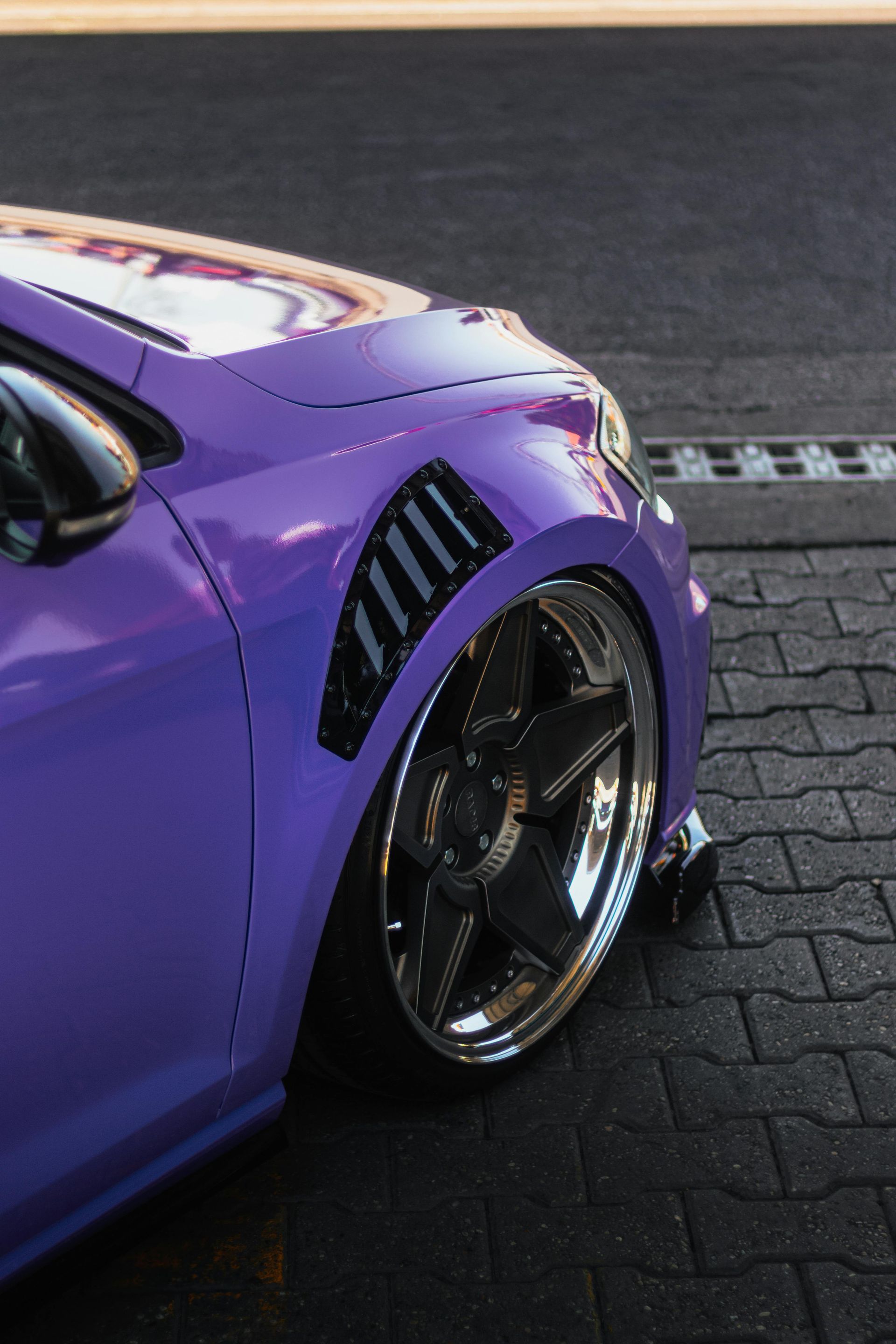 Purple car with chrome wheels, low to the ground, parked on a brick road. Side detail with vent.