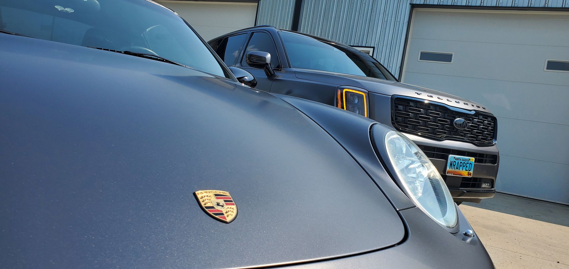 A dark gray Porsche sports car next to a dark SUV parked in front of a white garage.