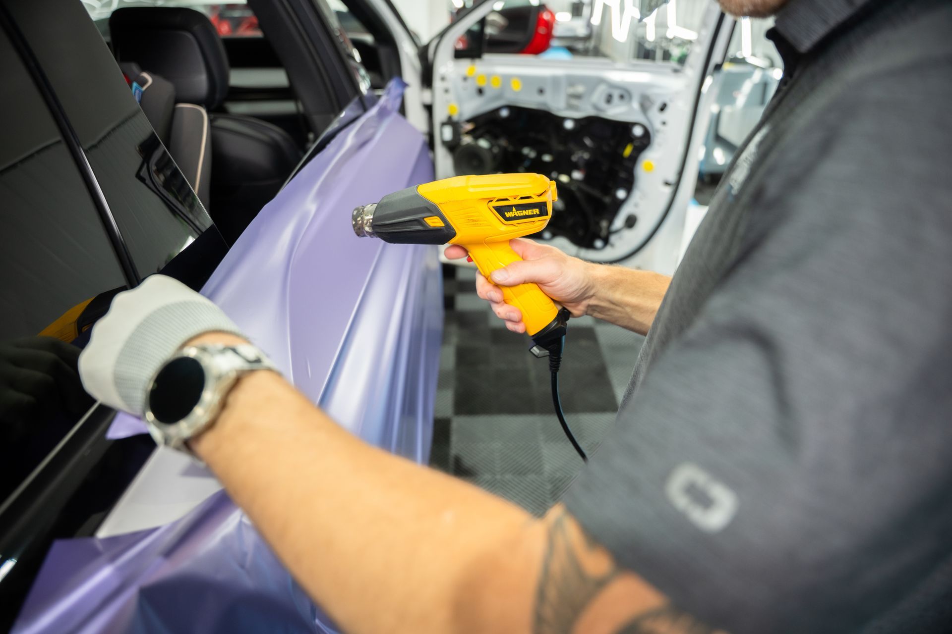 Person using a heat gun on car wrap. Lavender vinyl against white car door. Interior shop setting.