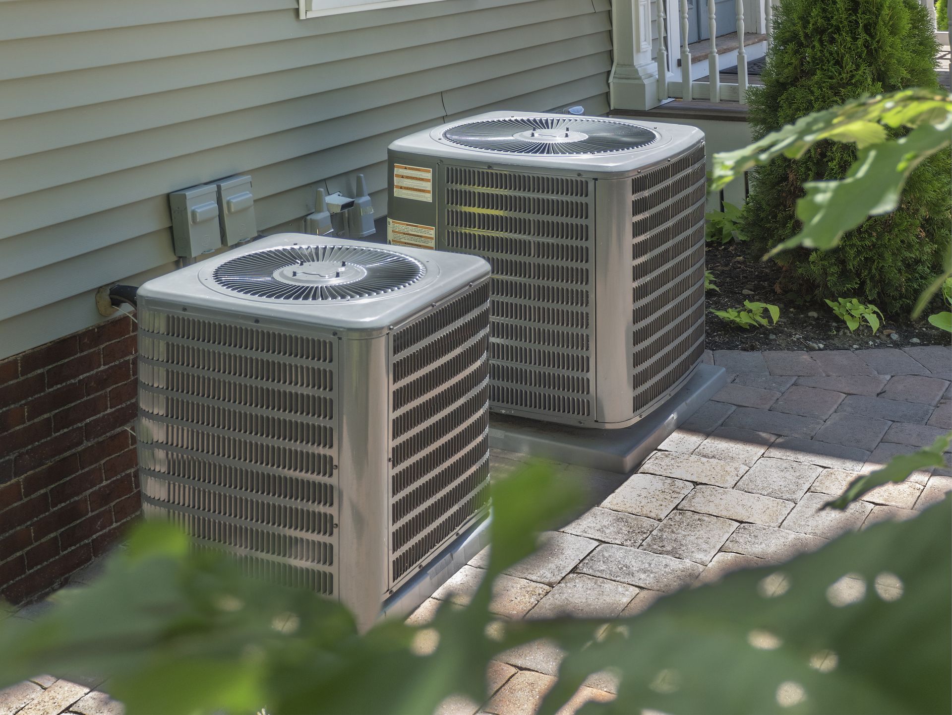Two heat pump units on a brick patio beside a house, green foliage in foreground.