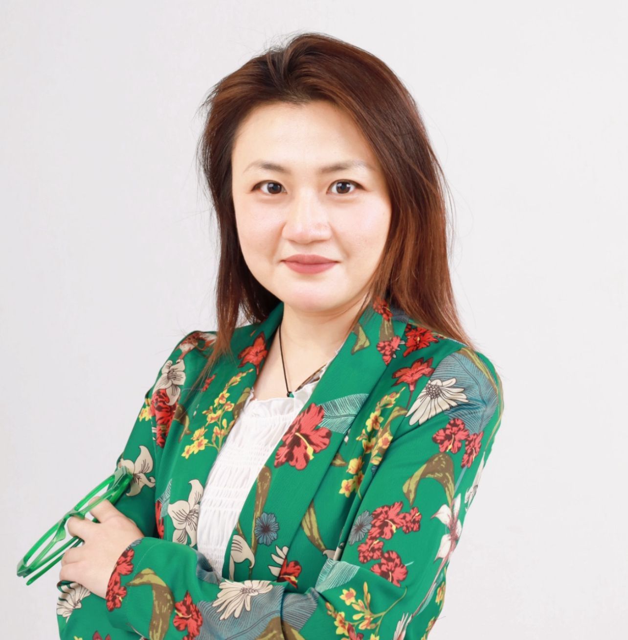 Woman in a green floral blazer with arms crossed, smiling at the camera. Light gray background.